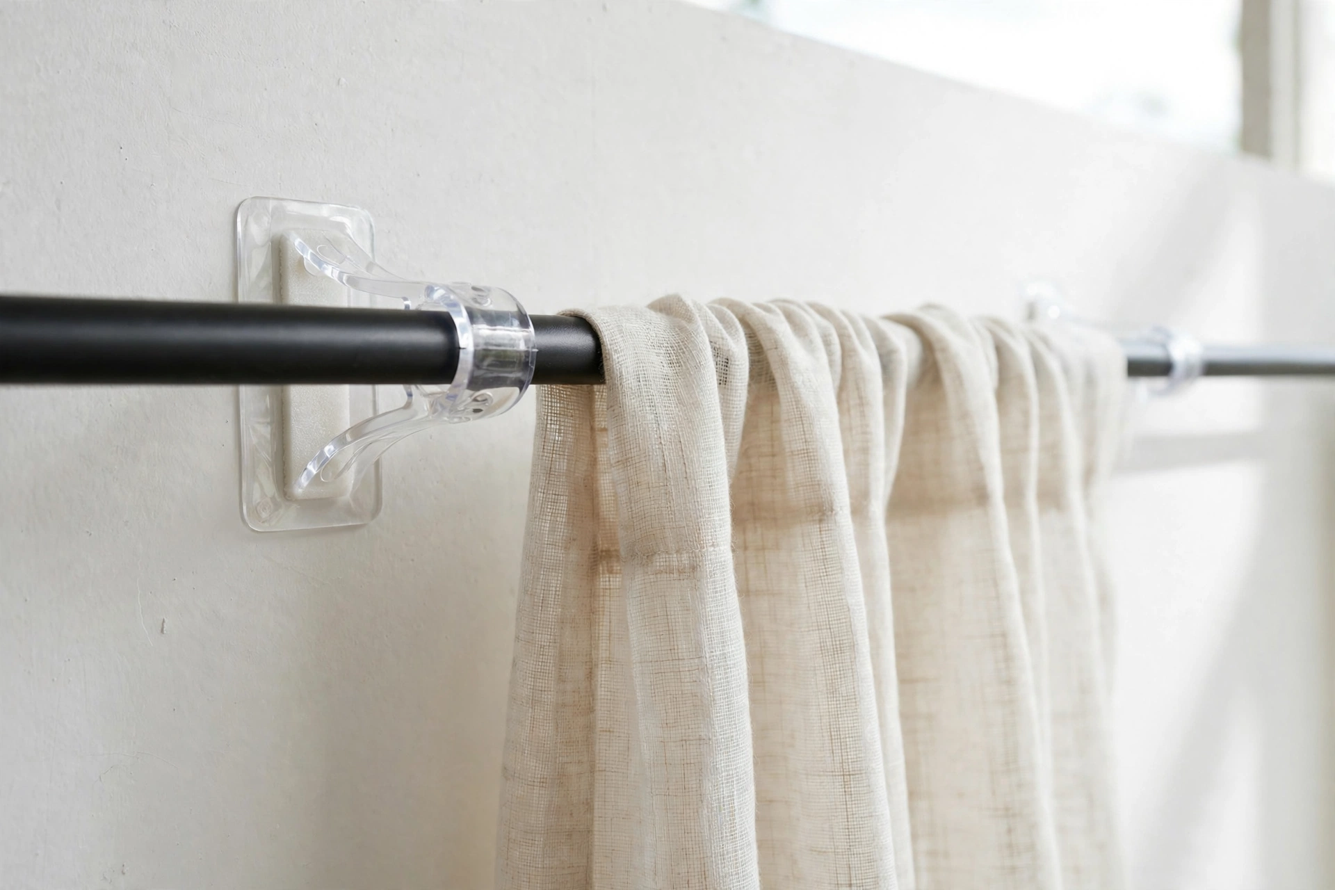 Close-up of clear adhesive curtain rod brackets holding a slim rod in a rental apartment